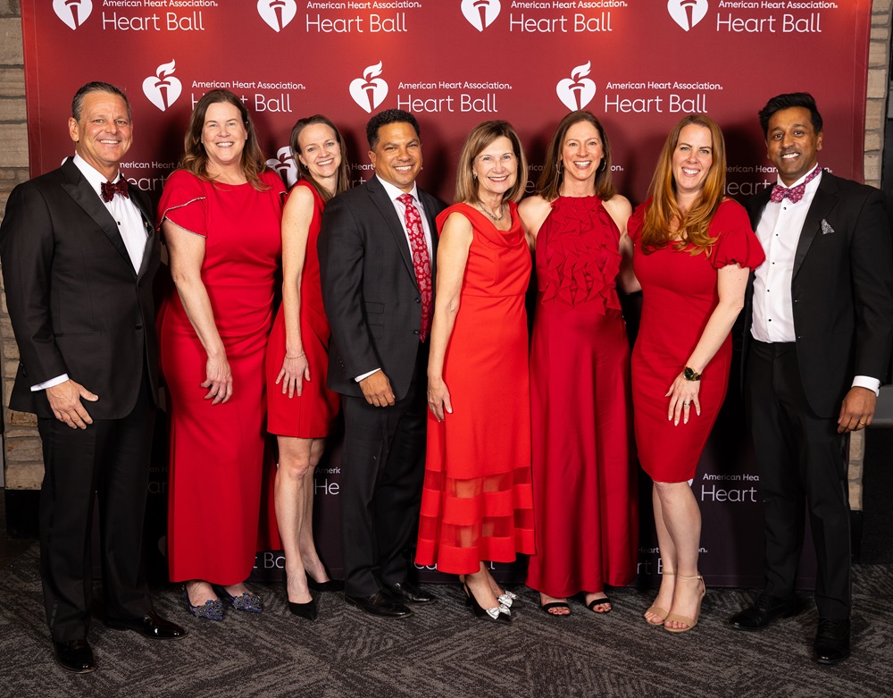 Group of eight people in formal dress standing in front of the American Heart Association Heart Ball step and repeat banner.